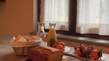 Un desayuno con pan, una botella de jugo de naranja y fruta fresca sobre una mesa cerca de una ventana con cortinas.