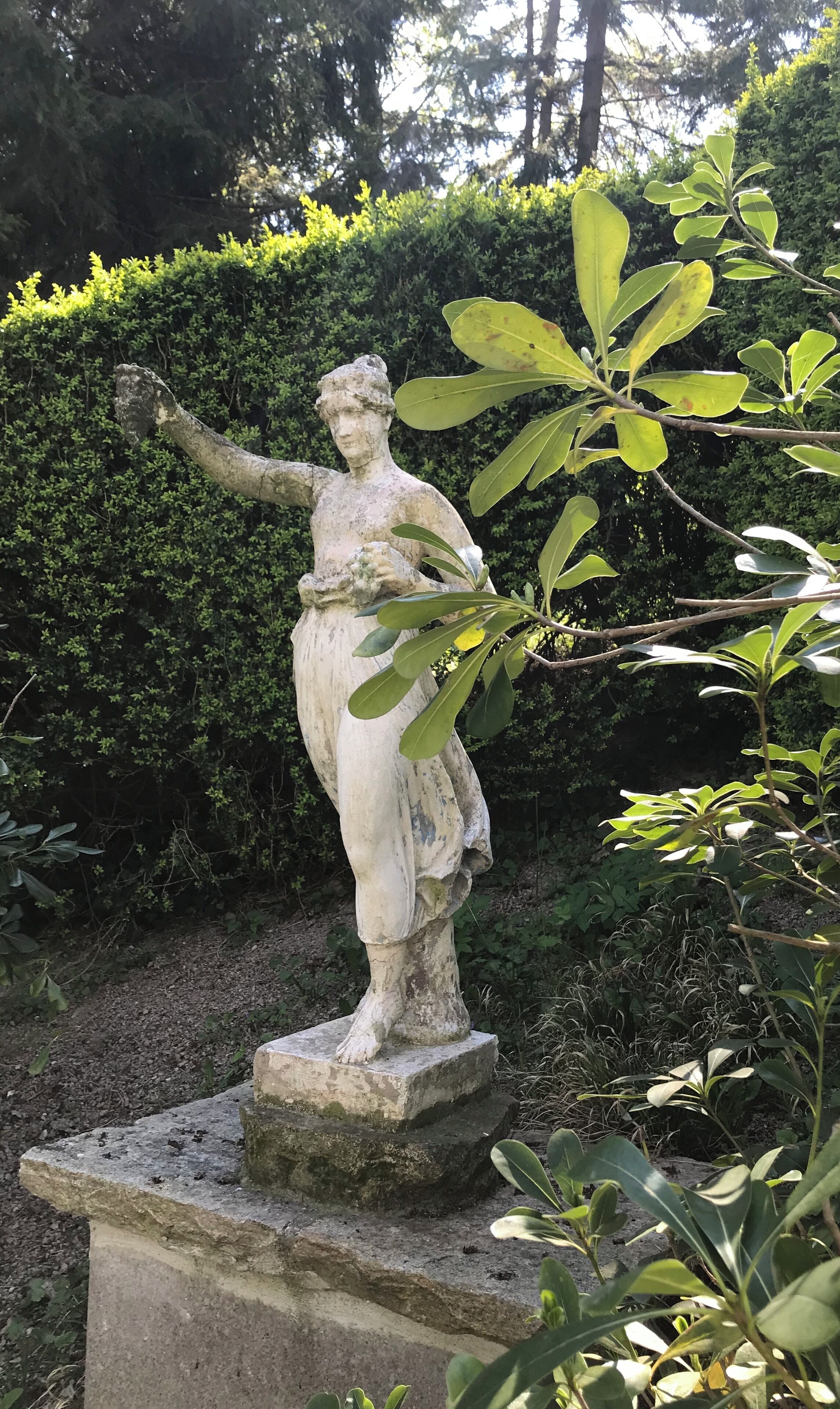 Una statua in pietra raffigura una figura con le braccia aperte, posata su un piedistallo in un giardino ricco di vegetazione.
