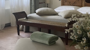 Un dormitori amb un llit, una cadira, una banca, una làmpada i una finestra amb cortines.