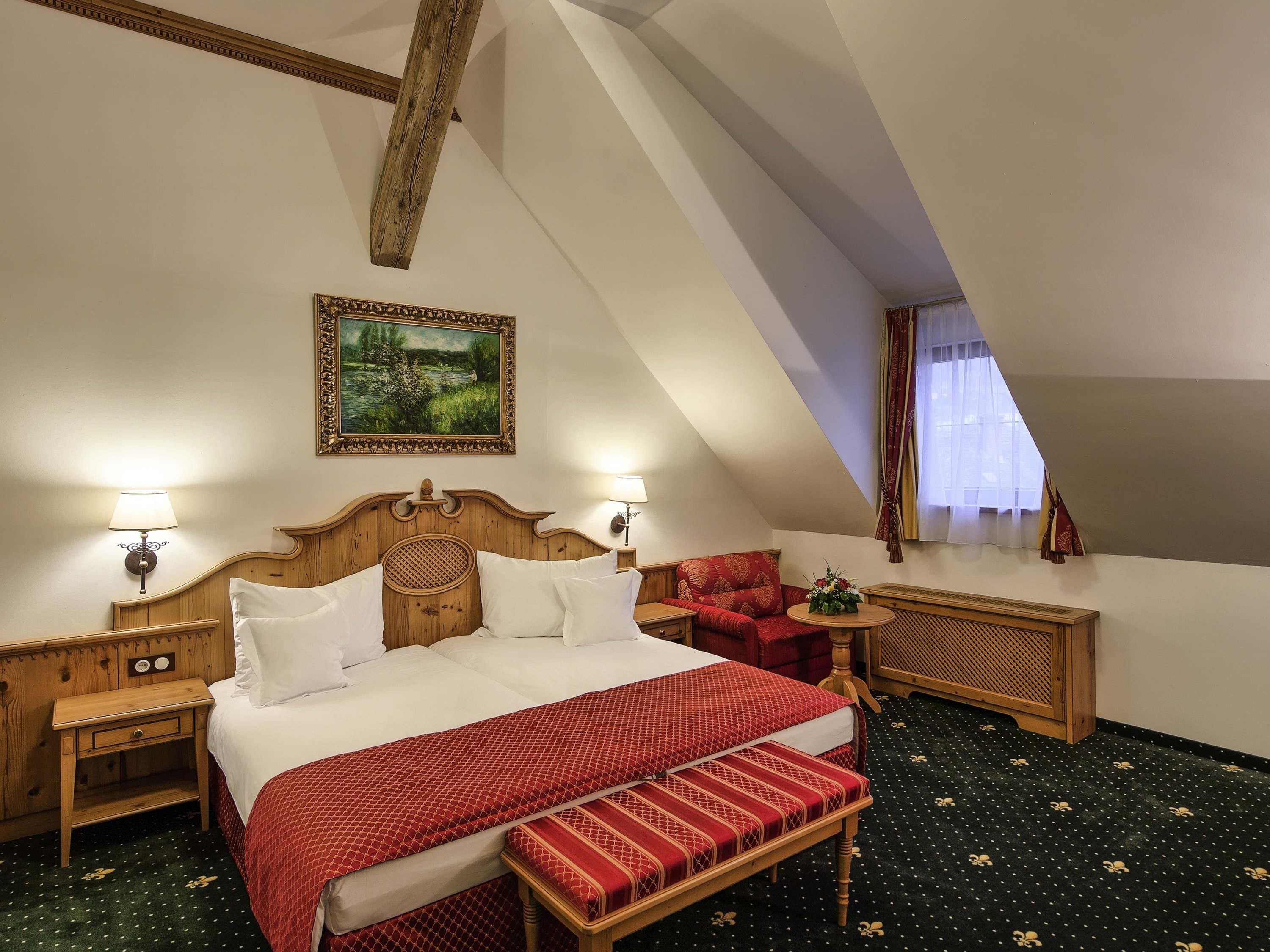 A bedroom with a large bed, two bedside tables, a red bench, a wooden headboard, a painting, and a window with curtains.