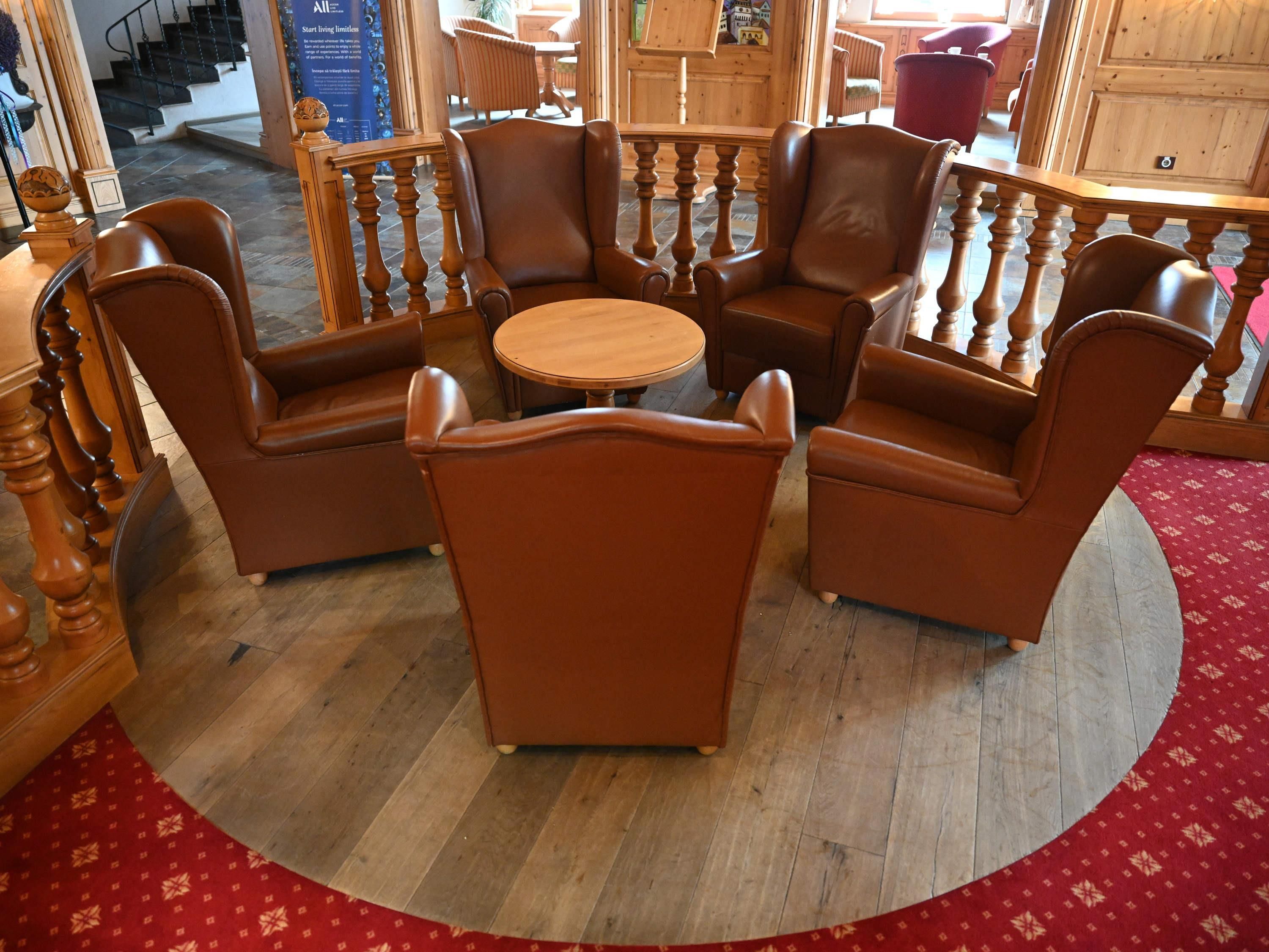A circular seating area with a wooden table surrounded by four brown leather chairs, a wooden railing, and a red patterned carpet.