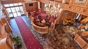 Un vaste hall d’entrée avec un plafond haut, des boiseries et un lustre. On y trouve un espace salon avec des canapés et des fauteuils, et un tapis rouge traverse l’ensemble de la pièce.