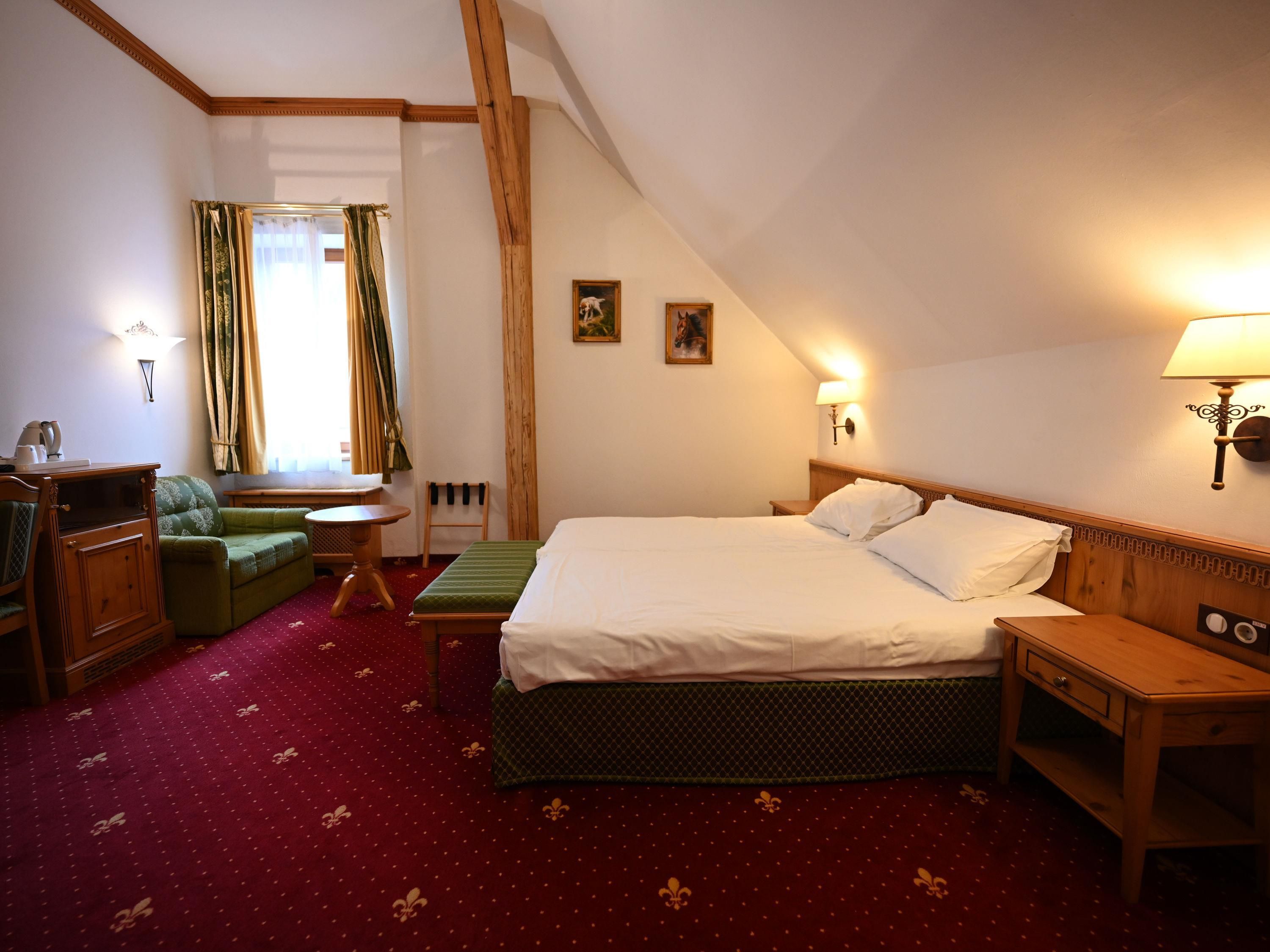 A hotel room with a bed, a green sofa, a wooden nightstand, a small table, and a window with curtains.