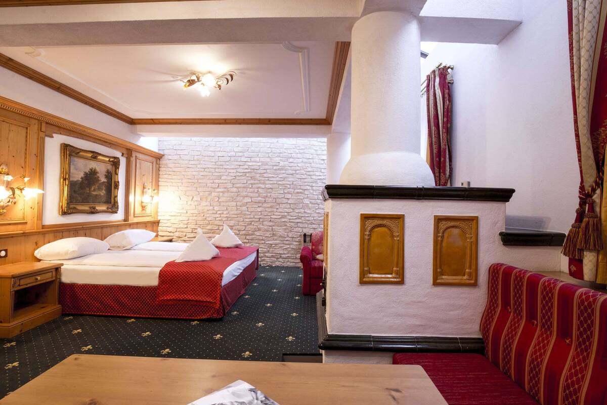 A hotel room with a bed, a wooden nightstand, a table, a red sofa, a stone wall, and a fireplace.