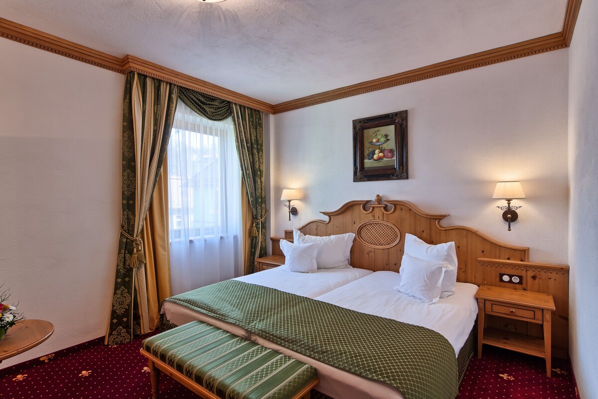 A hotel room with a large bed, two bedside tables with lamps, a wooden headboard, a framed picture on the wall, and a window with curtains.
