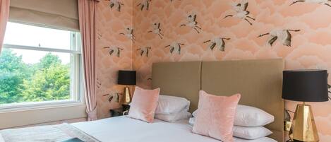 A bedroom with a large bed, two bedside tables with lamps, a window with curtains, and a wall with a bird pattern.