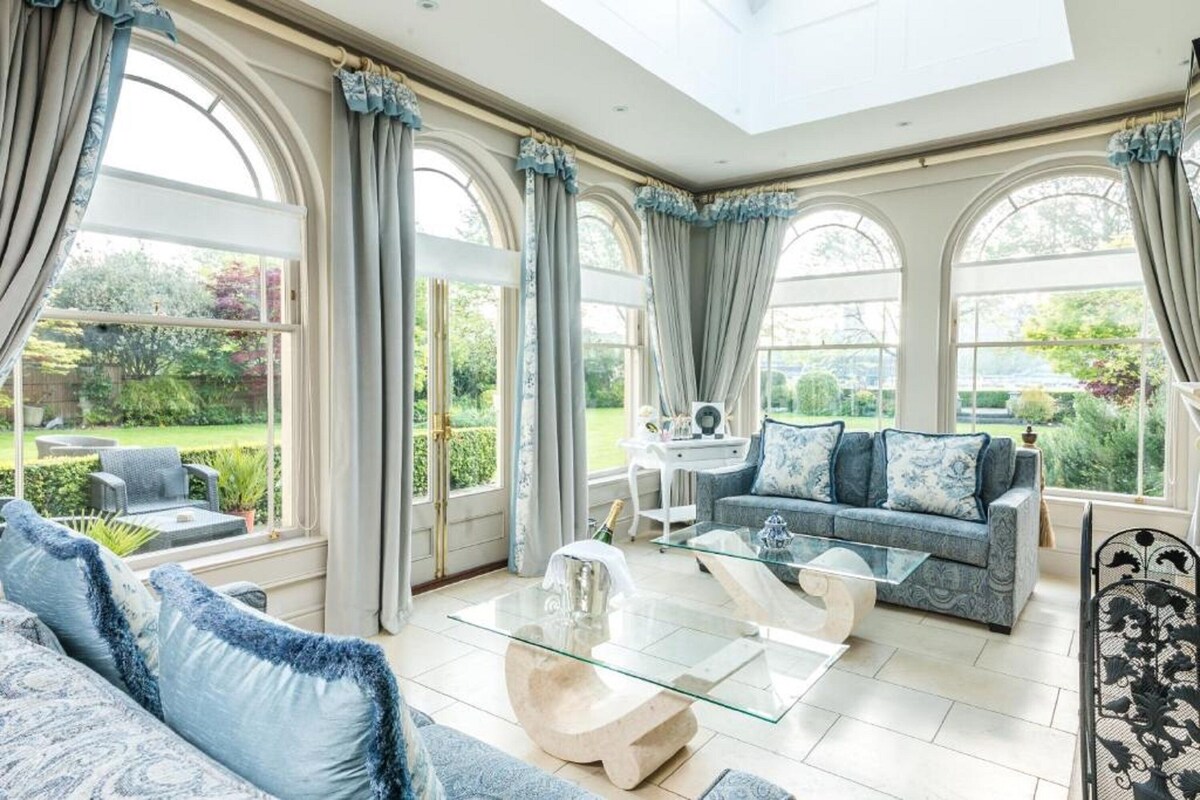 A spacious living room with a glass coffee table, a sofa, and a dining table. Large windows with curtains provide views of the garden.