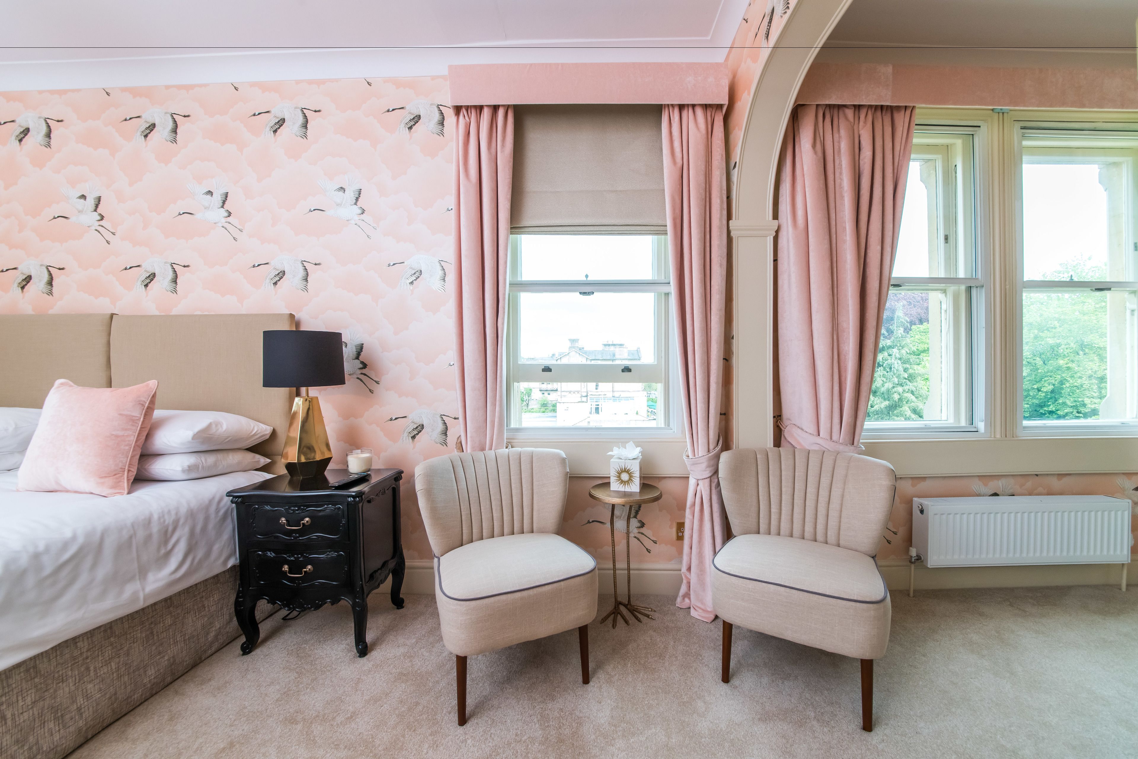 Une chambre à coucher comprenant un lit, deux fauteuils, une table de chevet, une lampe, une petite table et une fenêtre avec des rideaux.
