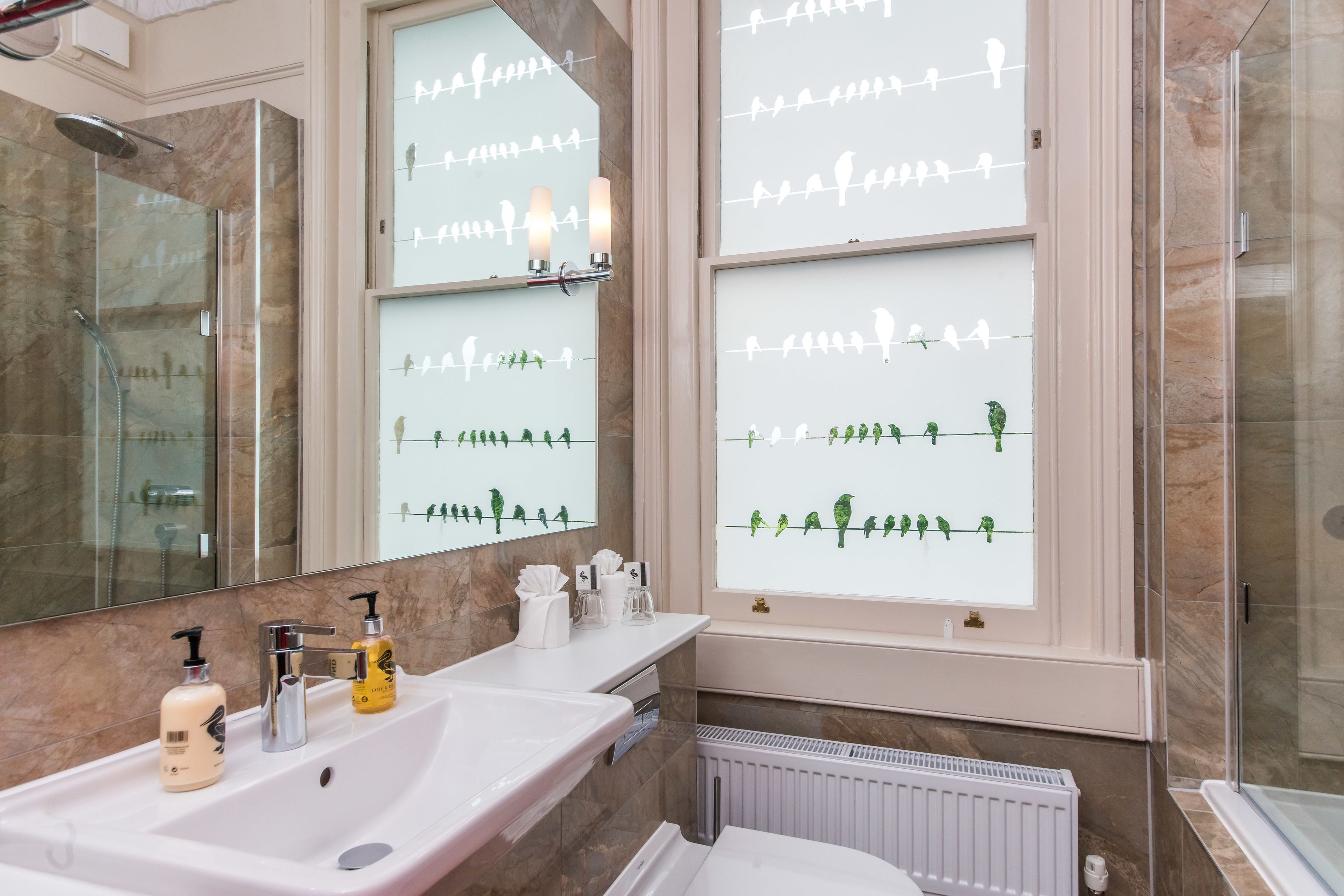 Een badkamer met een marmeren wand, een witte wastafel met twee zeepflessen, een spiegel en een raam met silhouetten van vogels.