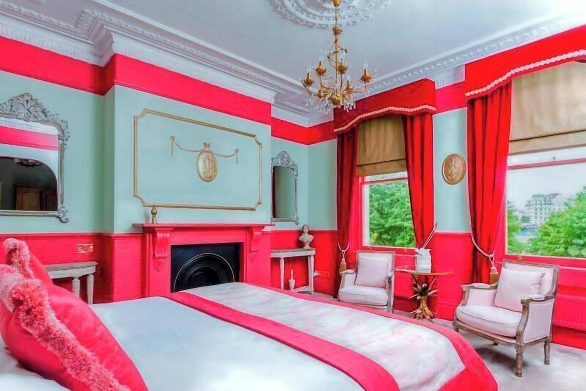 A room with a bed, a fireplace, two chairs, a chandelier, and a window with curtains.