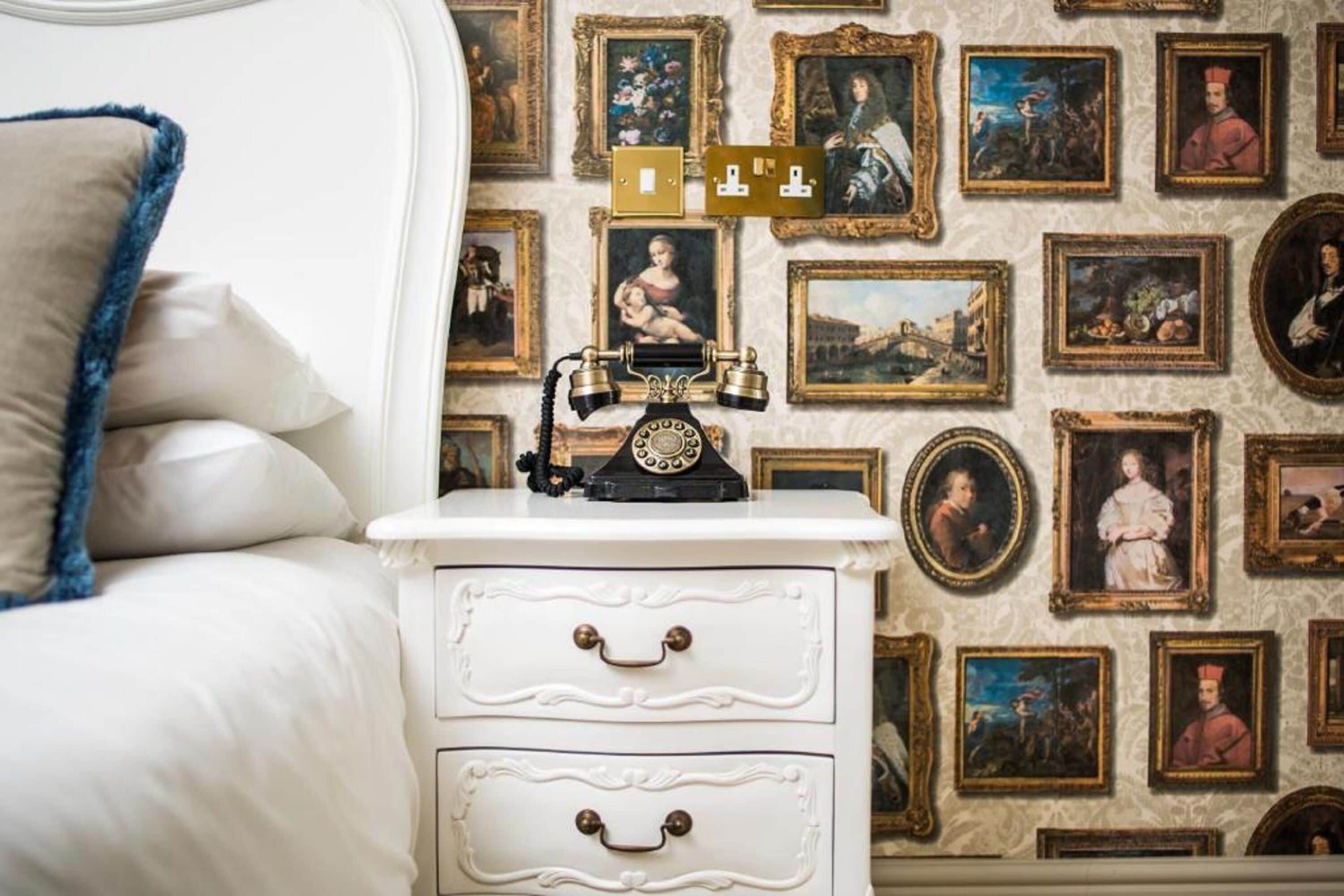 A bedroom with a white bed, a white nightstand with drawers, and a wall adorned with framed paintings.