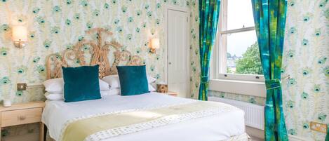 A bedroom with a bed, two bedside tables, a window with curtains, and wallpaper with a peacock feather pattern.