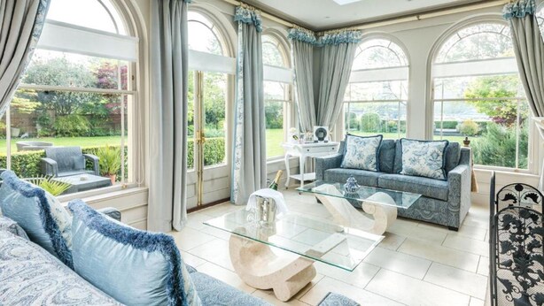 A spacious living room with a sofa, coffee table, and a glass-top table. There are large windows with curtains, a view of the garden, and a skylight. - The Ayrlington (Bath)