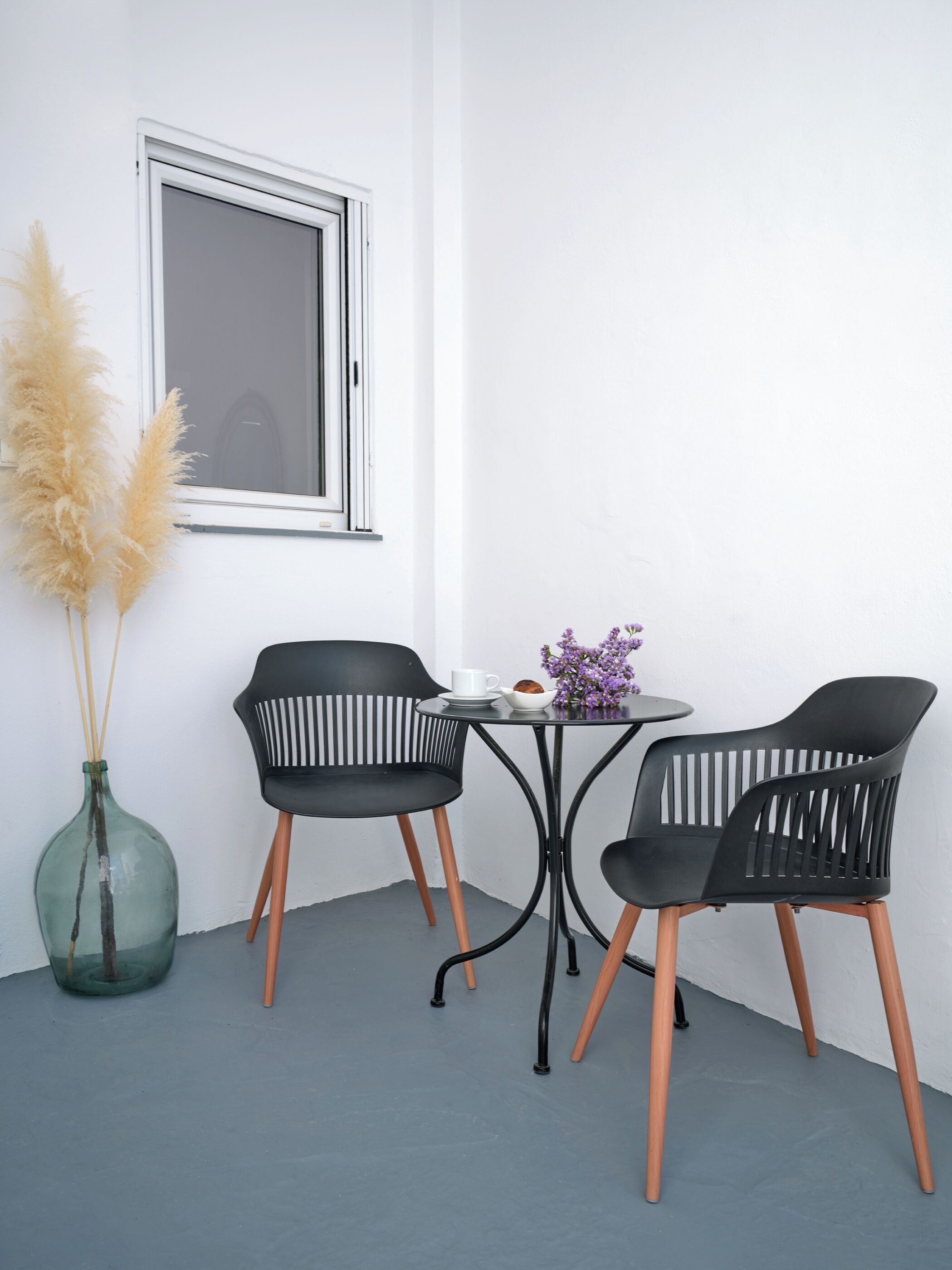 Una habitación pequeña y moderna con una mesa redonda y dos sillas negras con patas de madera. Hay una ventana con marco blanco, un jarrón verde con plumas de pampa, y una mesita con una taza blanca, un cuenco y flores moradas.