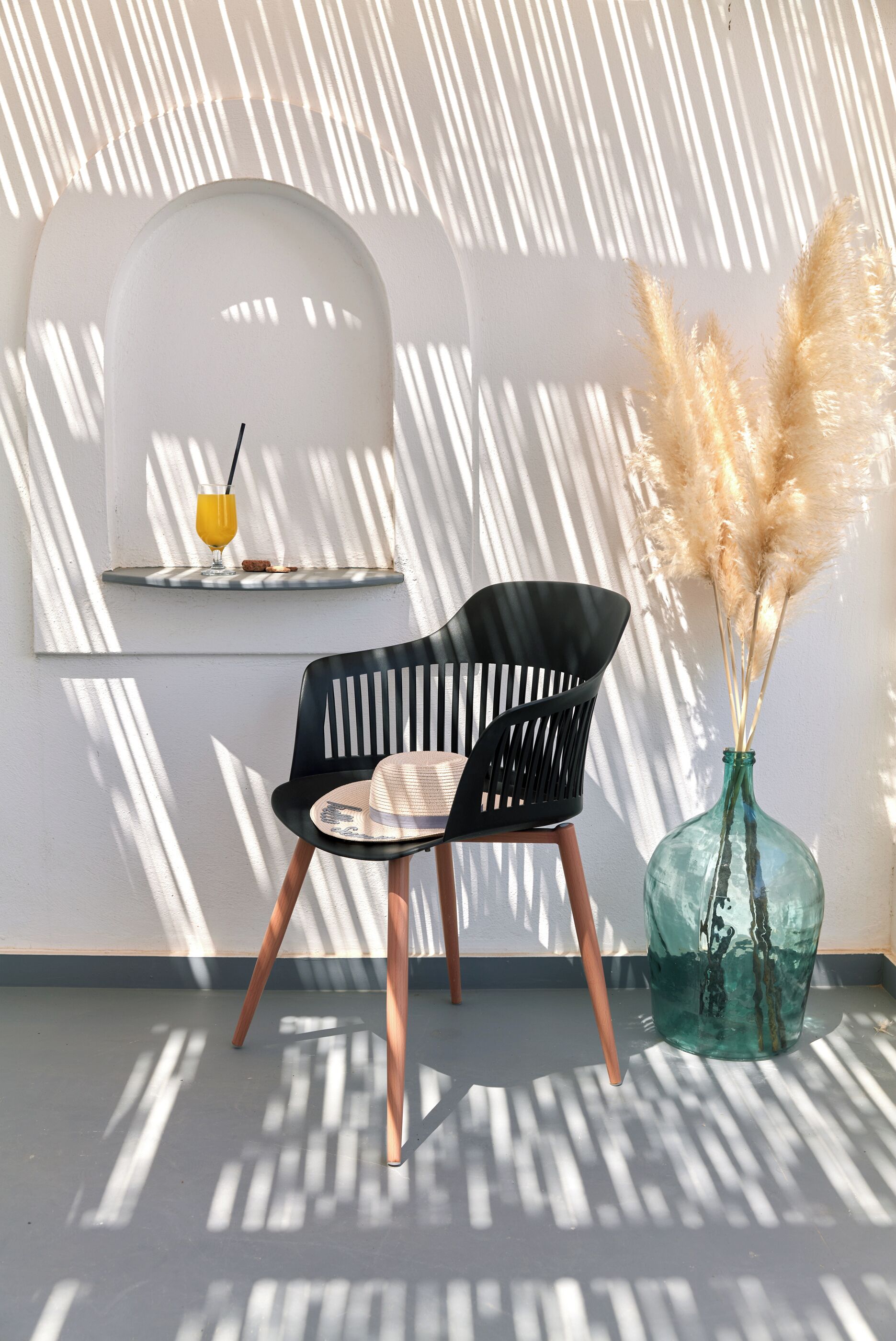 A modern room with a black chair, a glass vase with pampas grass, and a shelf with a glass of orange juice.