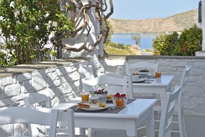 A table set with breakfast items, including fruit, yogurt, and juice, overlooking a scenic body of water. - Mersina Exclusive (Paros)