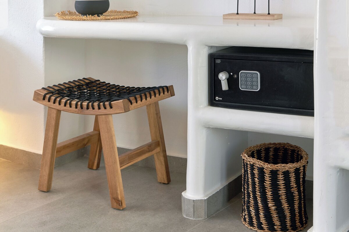 A white console table with a woven mirror, a small vase, and decorative leaves. A wooden stool is placed in front of the table.