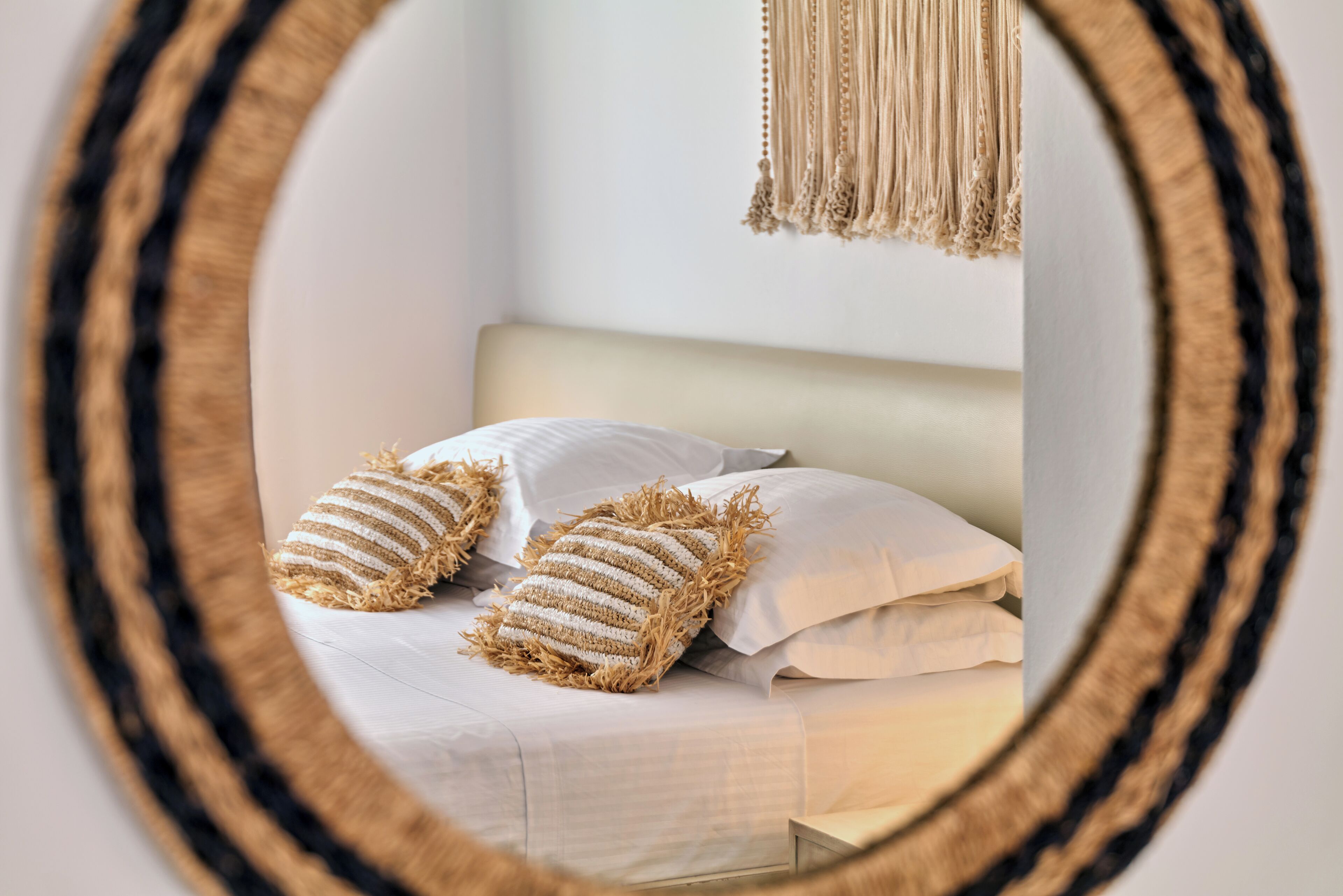 Una cama con sábanas blancas y dos almohadas a rayas, un adorno de macramé colgante y un marco de espejo circular.