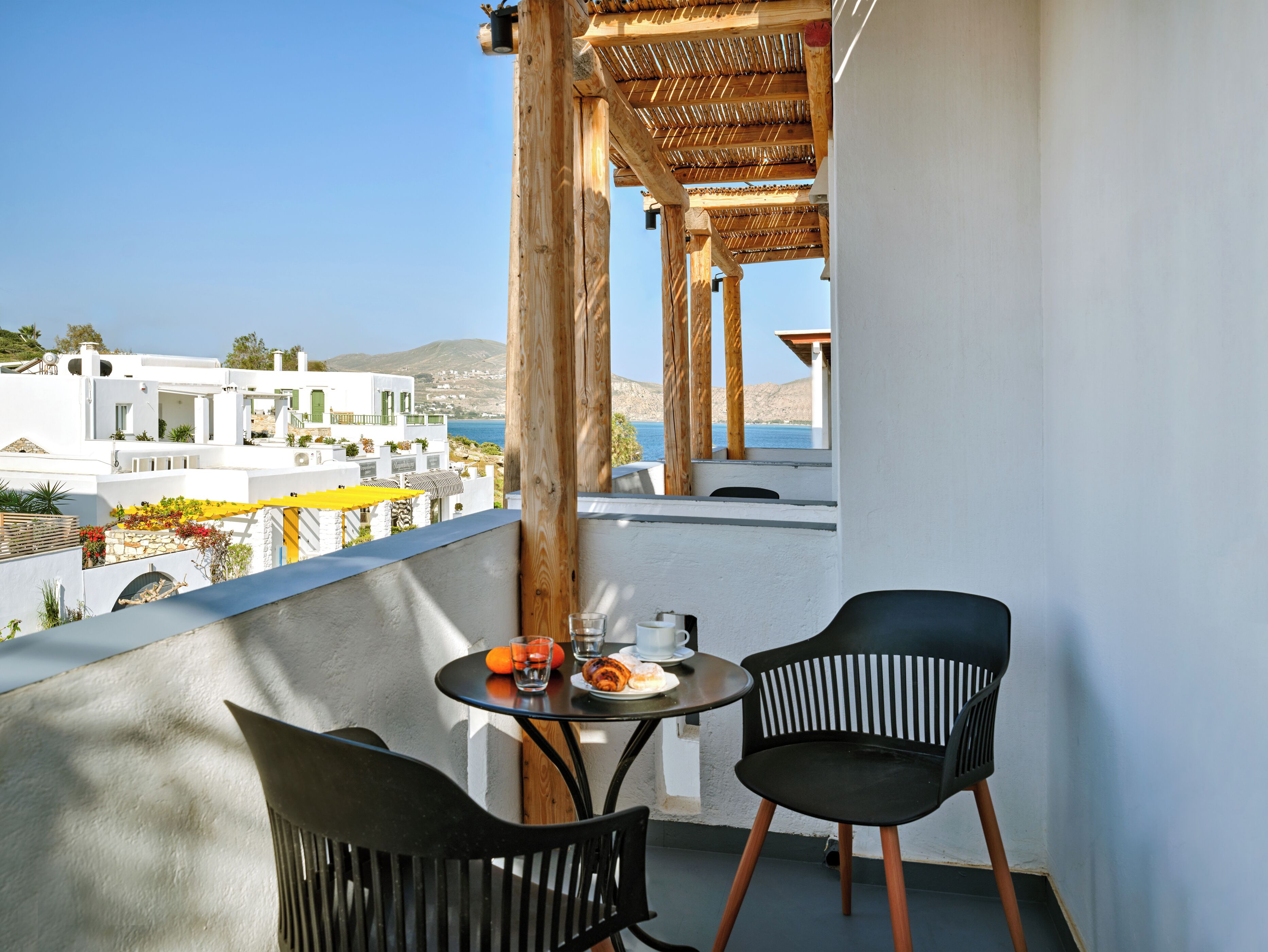 Un balcón con dos sillas negras, una mesa con comida y bebidas, y vistas a edificios blancos y al mar.