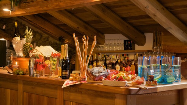 Zona de bar con vigas de madera, una variedad de botellas, vasos y una selección de frutas y aperitivos.