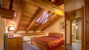 Un dormitorio acogedor en el ático con techo de madera, una cama, un escritorio con lámpara y un baño visible a través de una puerta abierta.