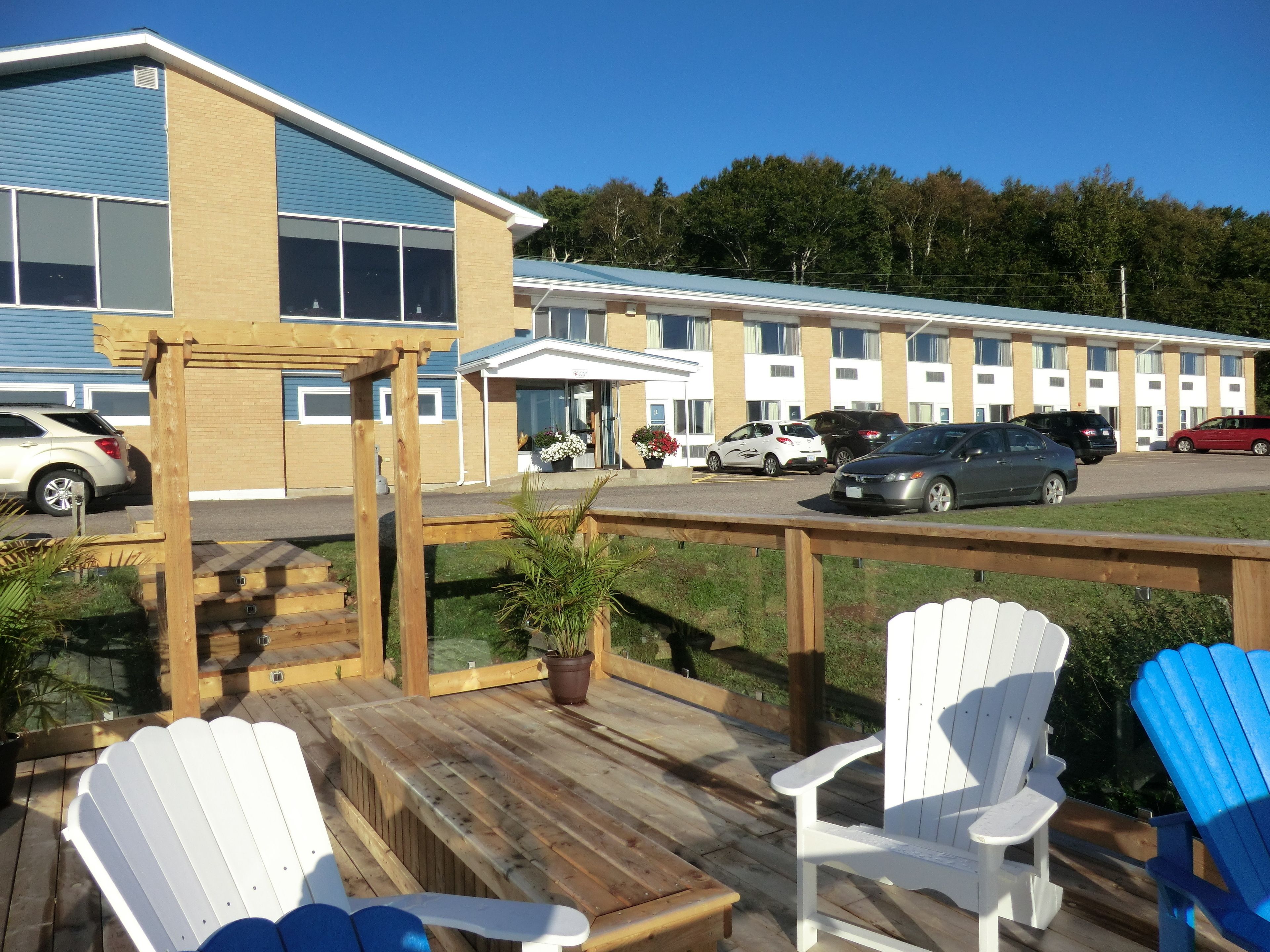 A motel with a deck area featuring white and blue Adirondack chairs, a covered patio, and a parking lot with several cars.