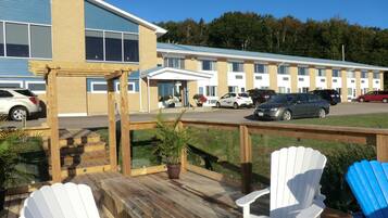 A motel with a deck area featuring white and blue Adirondack chairs, a covered patio, and a parking lot with several cars.