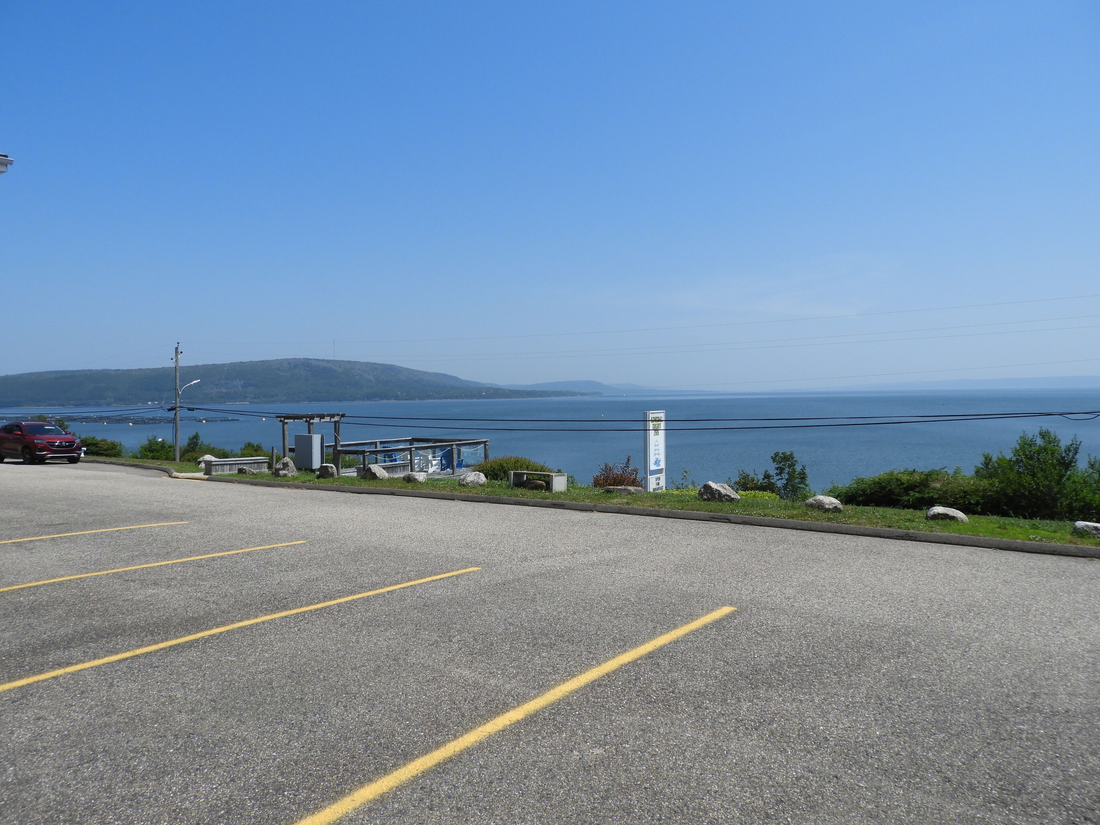 A parking lot with yellow lines, a red car, and a view of the sea and hills in the background.