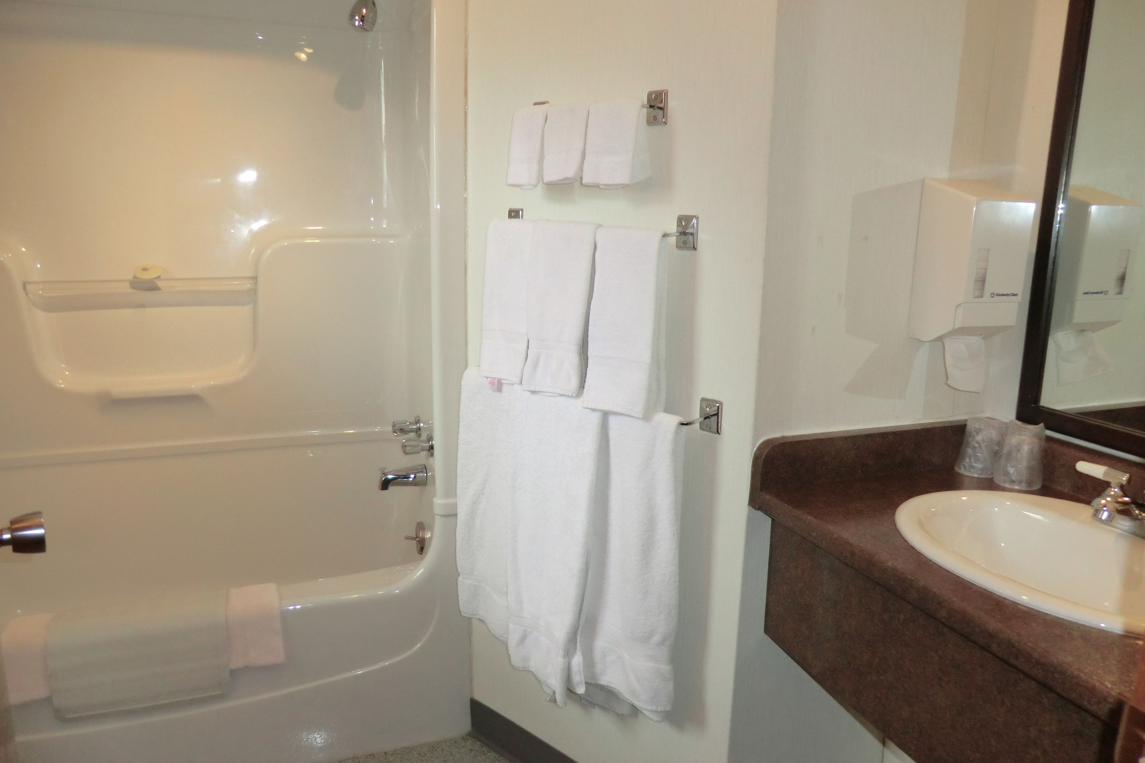 A bathroom with a bathtub, a sink, and a mirror. There are towels hanging on a rack and a soap dispenser on the wall.