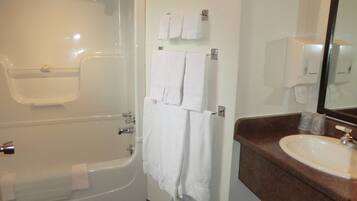 A bathroom with a bathtub, a sink, and a mirror. There are towels hanging on a rack and a soap dispenser on the wall.
