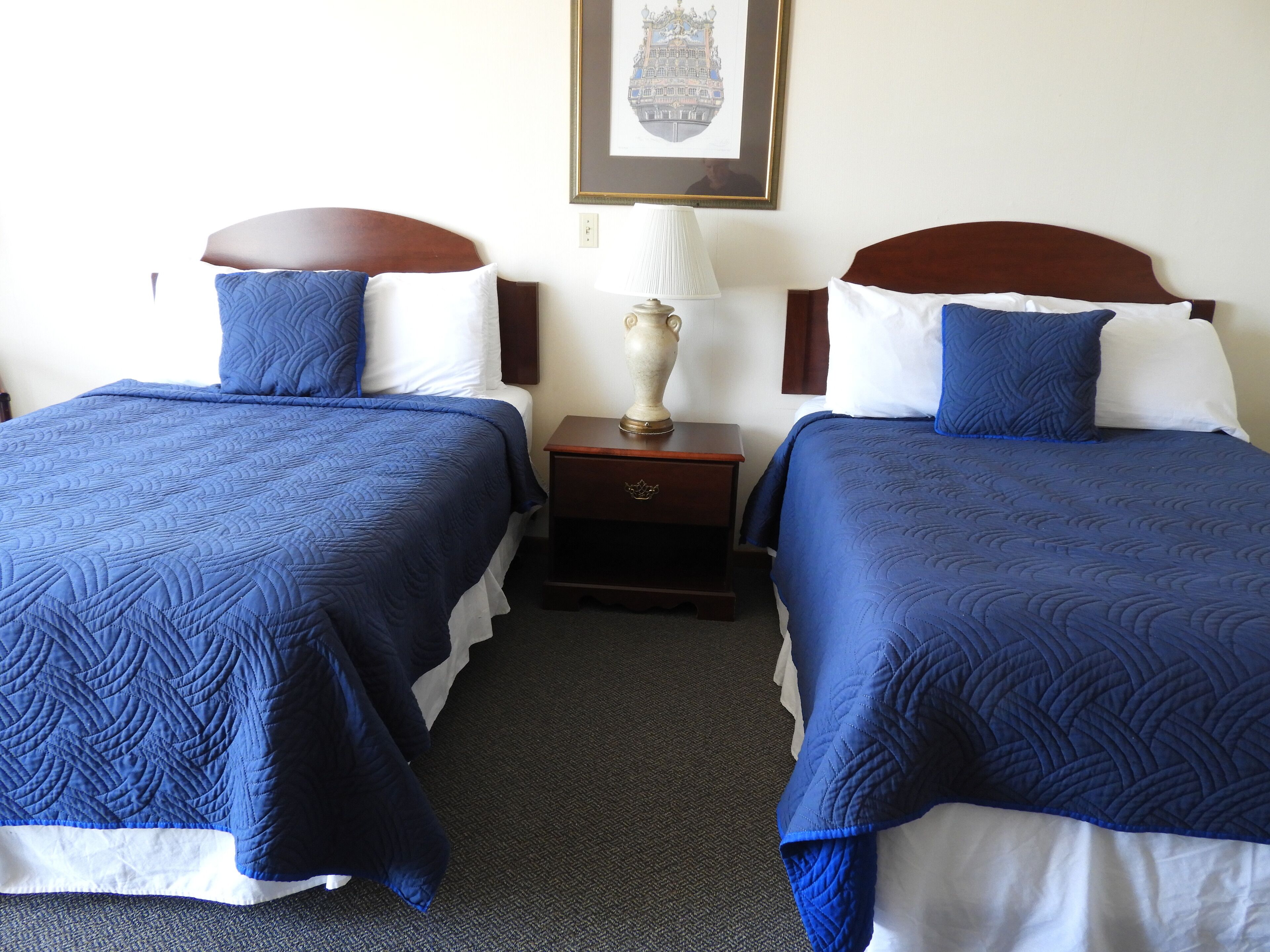 A hotel room with two beds, each with blue bedding and white pillows. There is a nightstand between the beds with a lamp and a small decorative item.