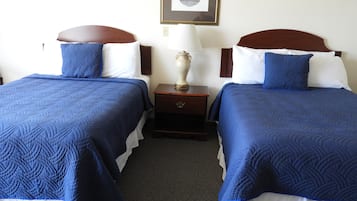 A hotel room with two beds, each with blue bedding and white pillows. There is a nightstand between the beds with a lamp and a small decorative item.
