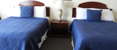 Habitación de hotel con dos camas, cada una con ropa de cama azul y almohadas blancas. Hay una mesita de noche entre las camas con una lámpara y un pequeño adorno.