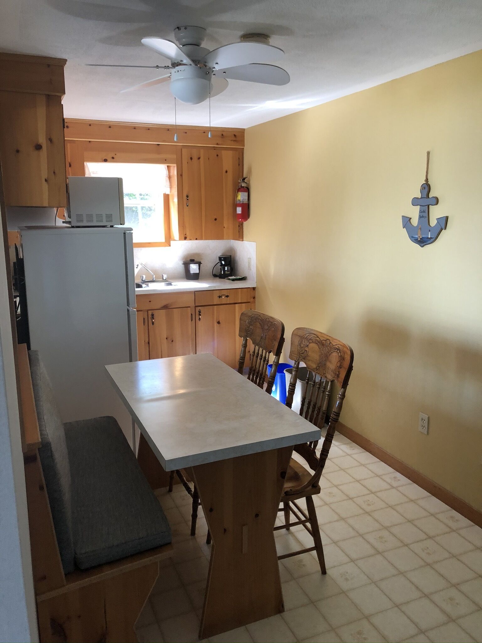 A compact kitchen with wooden cabinets, a built-in refrigerator, a dining table with chairs, a ceiling fan, and a wall-mounted fire extinguisher.