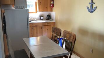 A compact kitchen with wooden cabinets, a built-in refrigerator, a dining table with chairs, a ceiling fan, and a wall-mounted fire extinguisher.