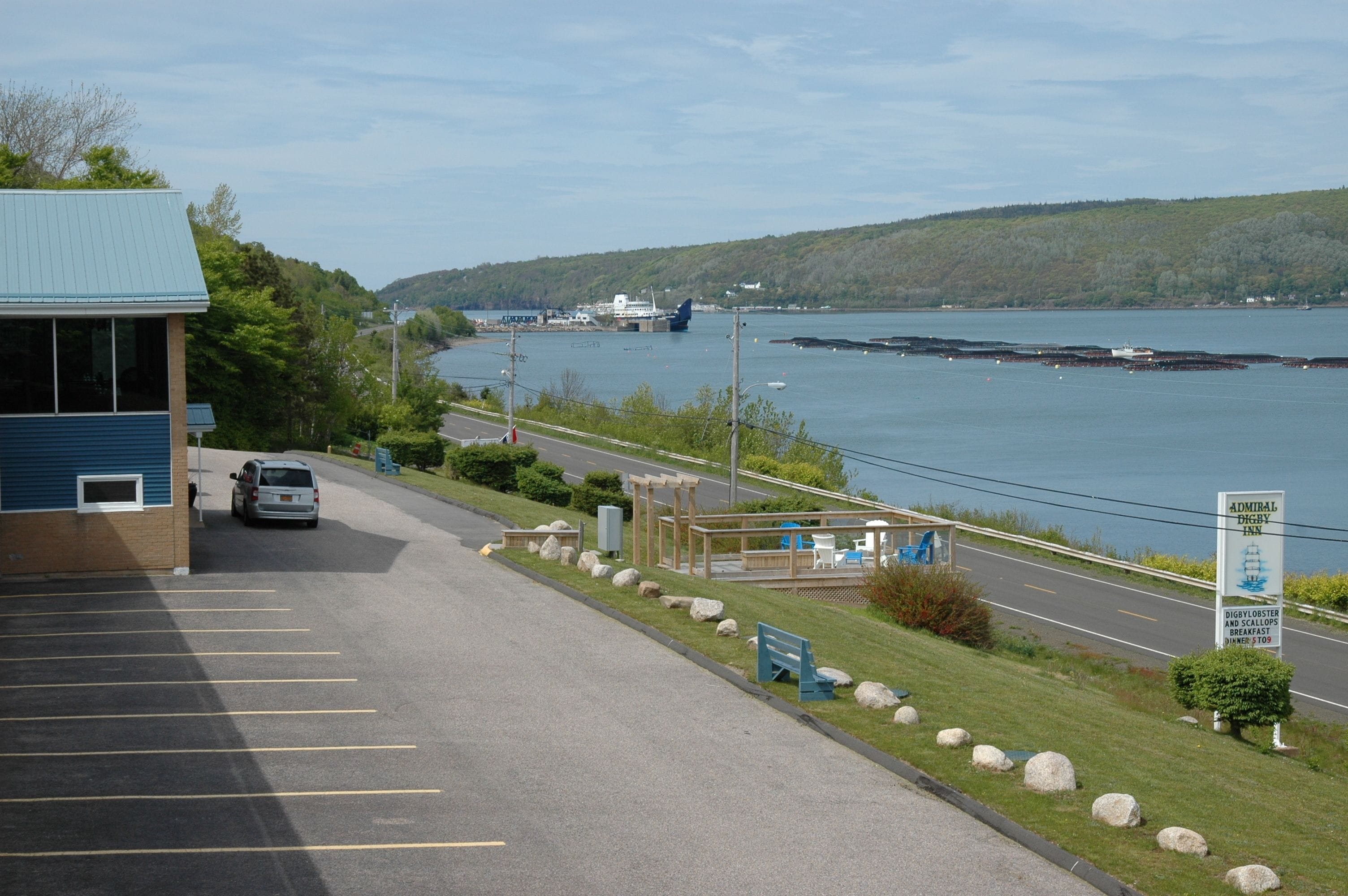 A parking lot with a building on the left, a wooden deck with seating, and a view of a body of water with a boat in the distance.