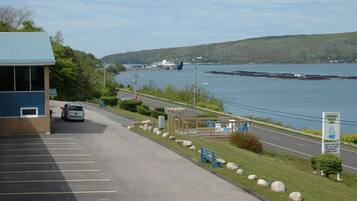 A parking lot with a building on the left, a wooden deck with seating, and a view of a body of water with a boat in the distance.