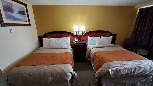 A hotel room with two beds, a nightstand with lamps, and a painting on the wall. - Imperial Fireside Inn (Sudbury)