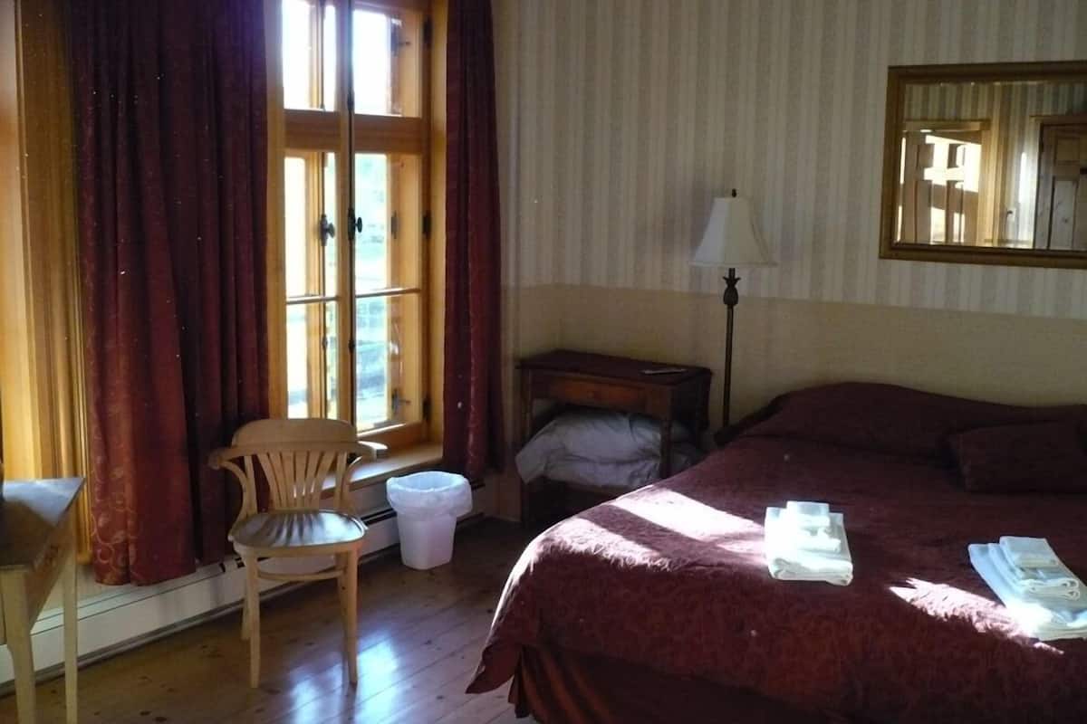 Hotellihuone, jossa on suuri sänky, tuoli, pieni pöytä, lamppu ja ikkuna, jossa on verhot.