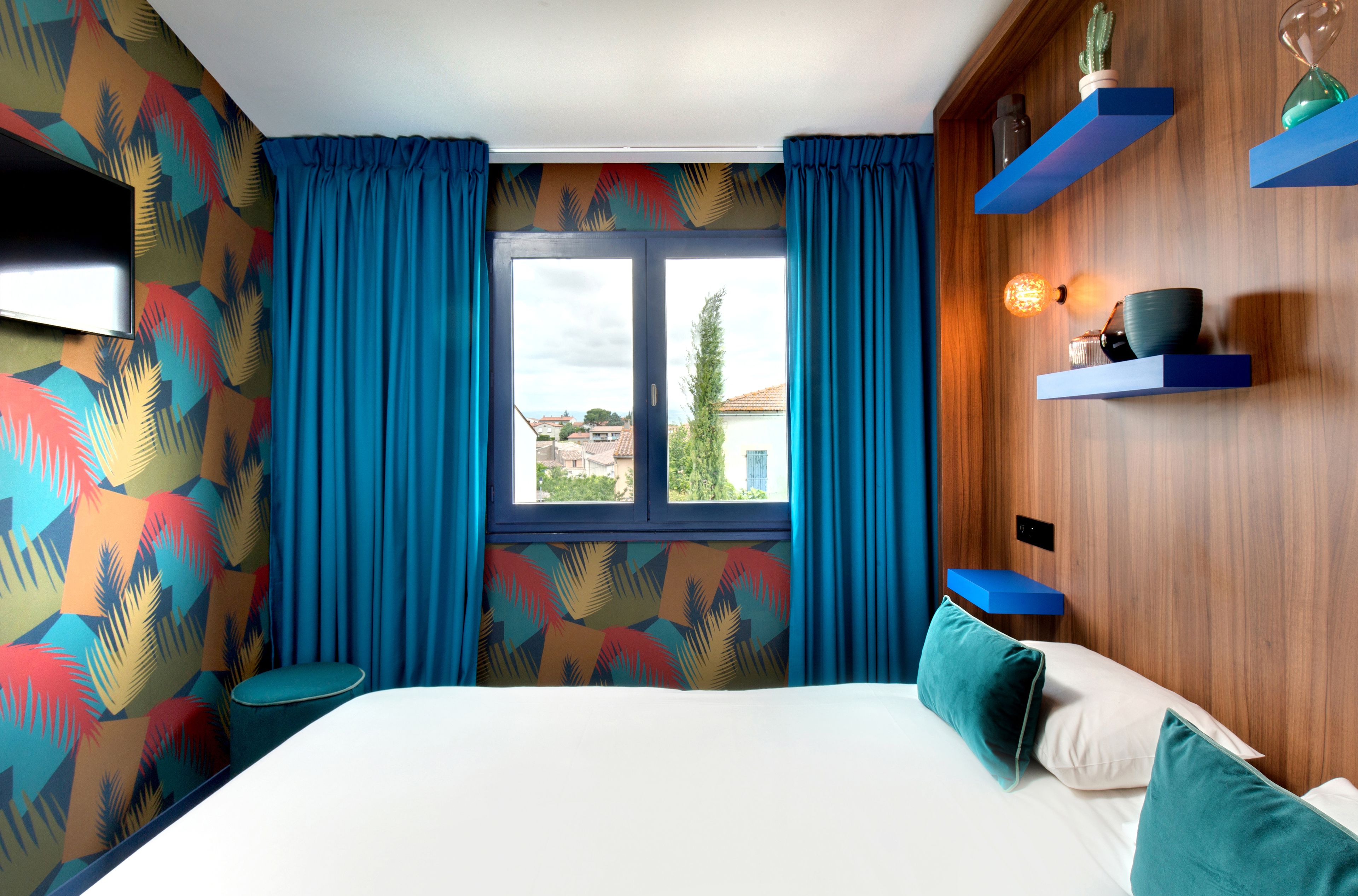 Een slaapkamer met een bed, gordijnen, een televisie en een raam met uitzicht op gebouwen.