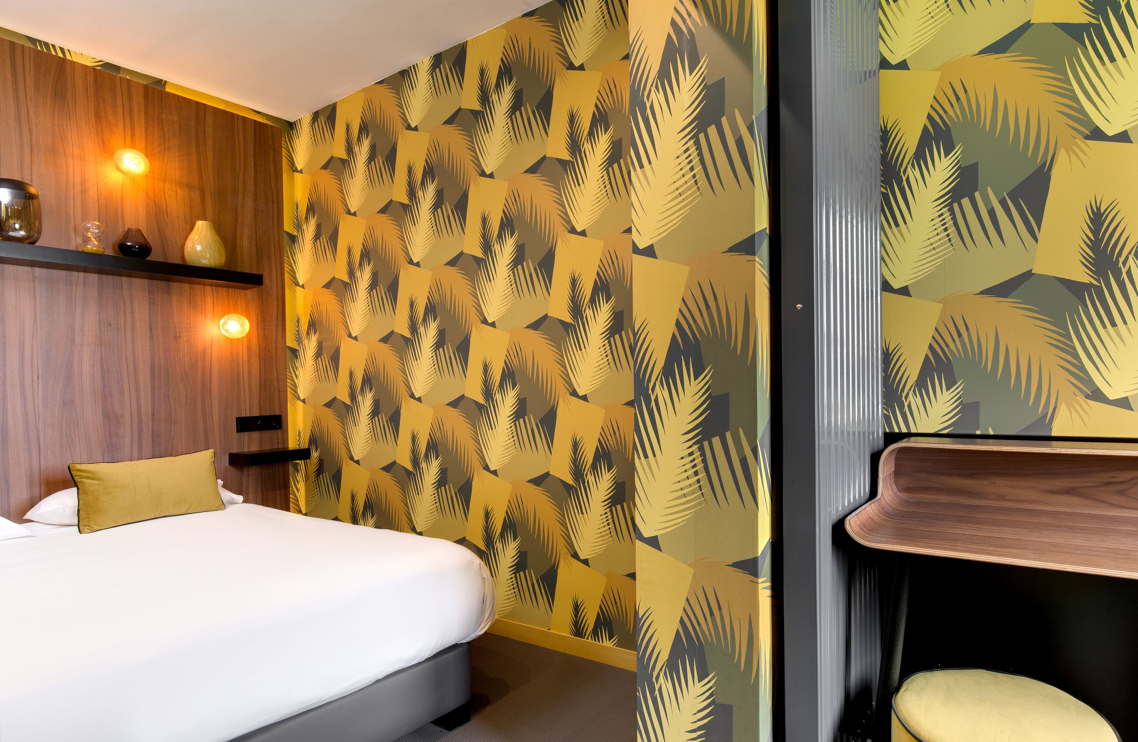 A hotel room with a bed, a wooden shelf with decorative items, and a patterned wallpaper.