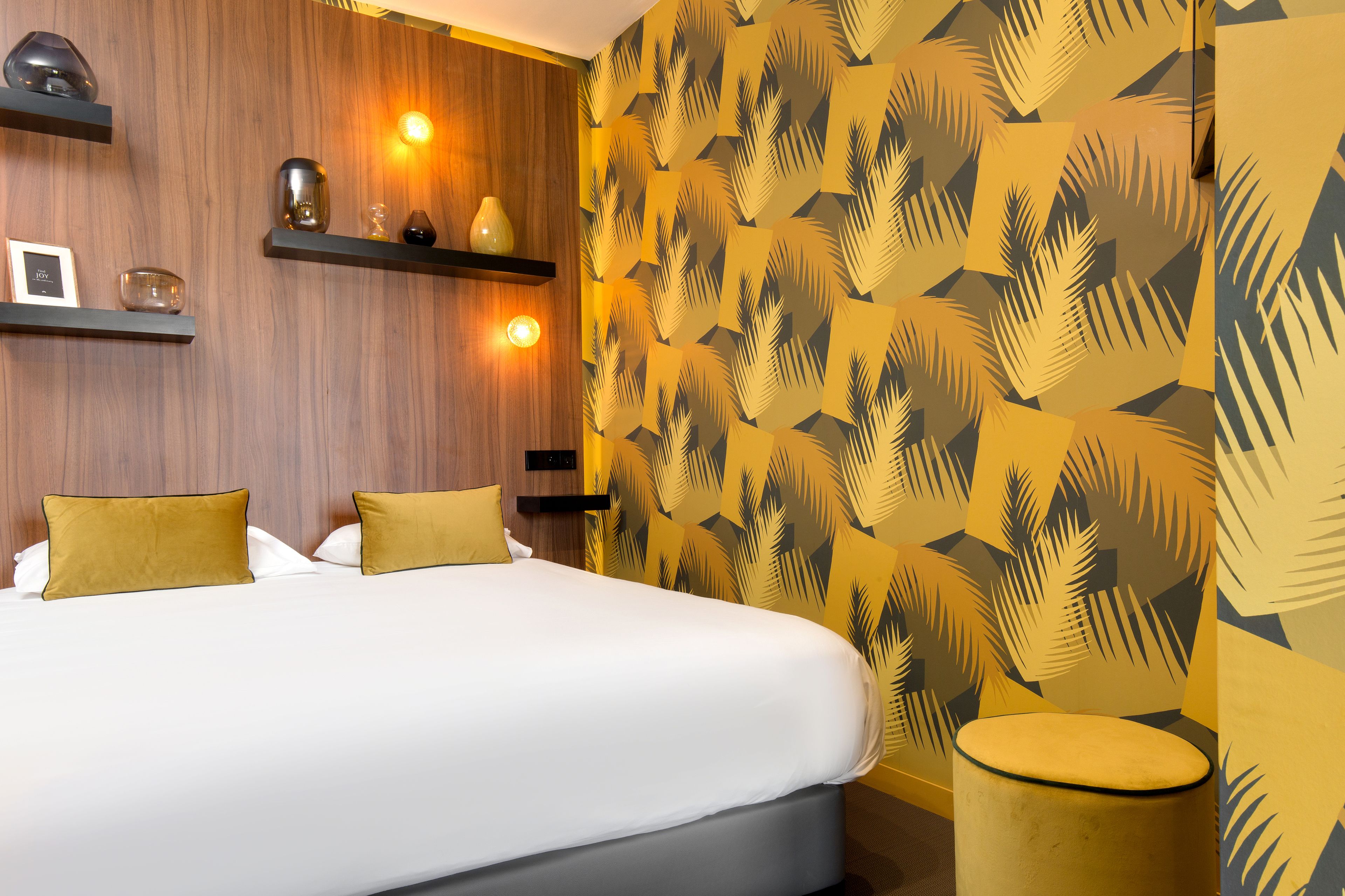 Ein Hotelzimmer mit einem Bett, zwei Kissen, einem Nachttisch mit Lampe und einer Wand mit einem Blattmuster.