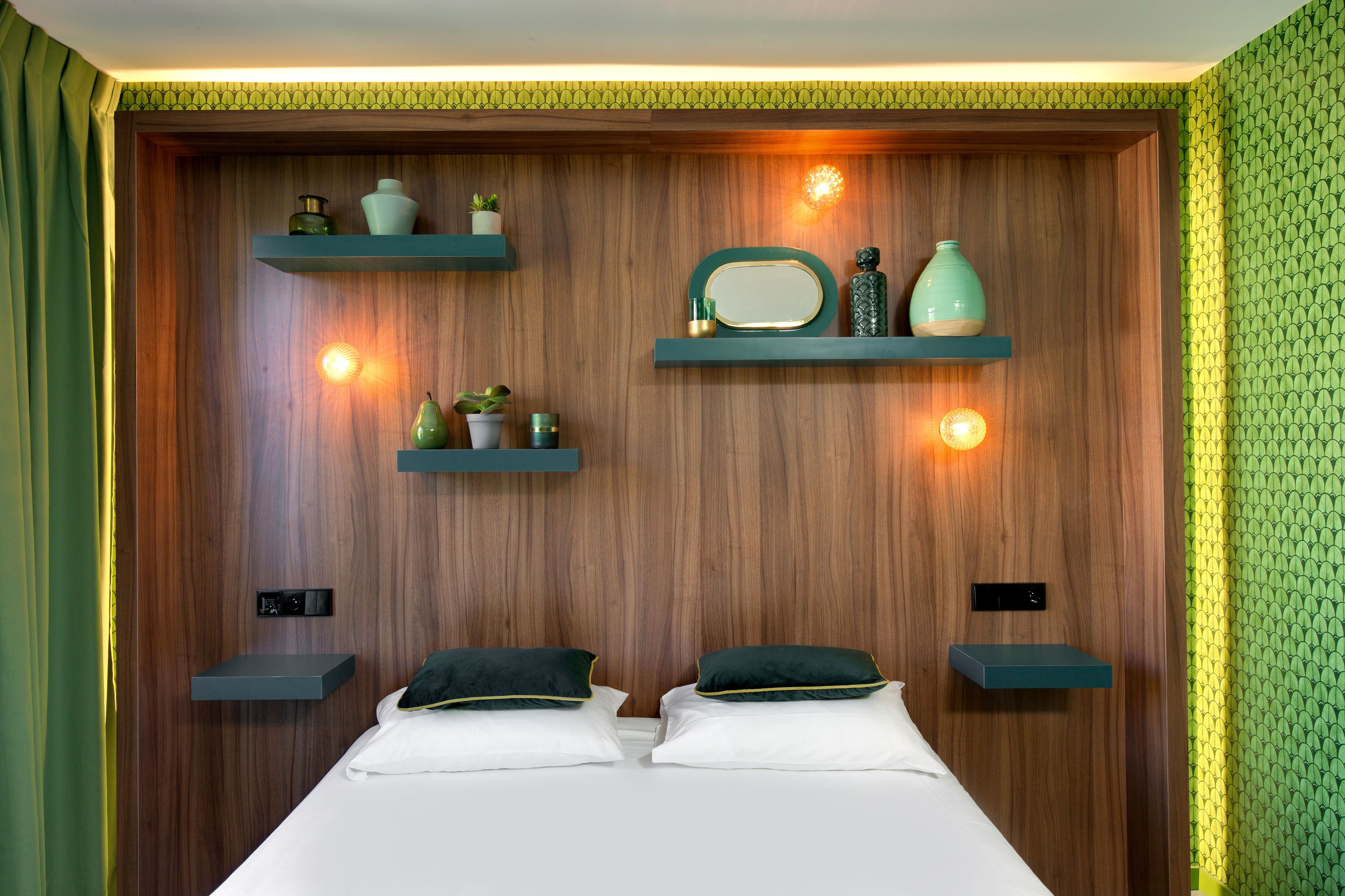 A bedroom with a wooden headboard, two bedside tables, a bed with white linens and dark green pillows, and wall-mounted shelves with decorative items.