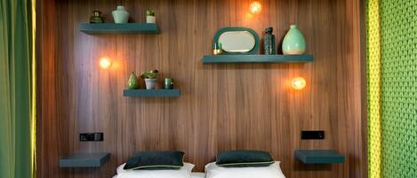 A bedroom with a wooden headboard, two bedside tables, a bed with white linens and dark green pillows, and wall-mounted shelves with decorative items.
