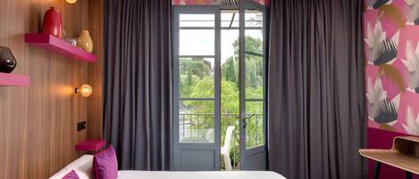Habitación de hotel moderna con una cama, una silla, una mesita y vistas a zonas verdes a través de una puerta.