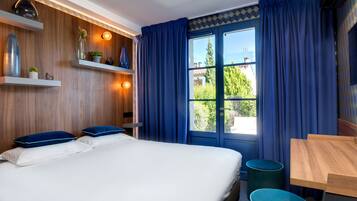 A modern hotel room with a large bed, a wooden wall with shelves, a window with blue curtains, and a small table with a chair.