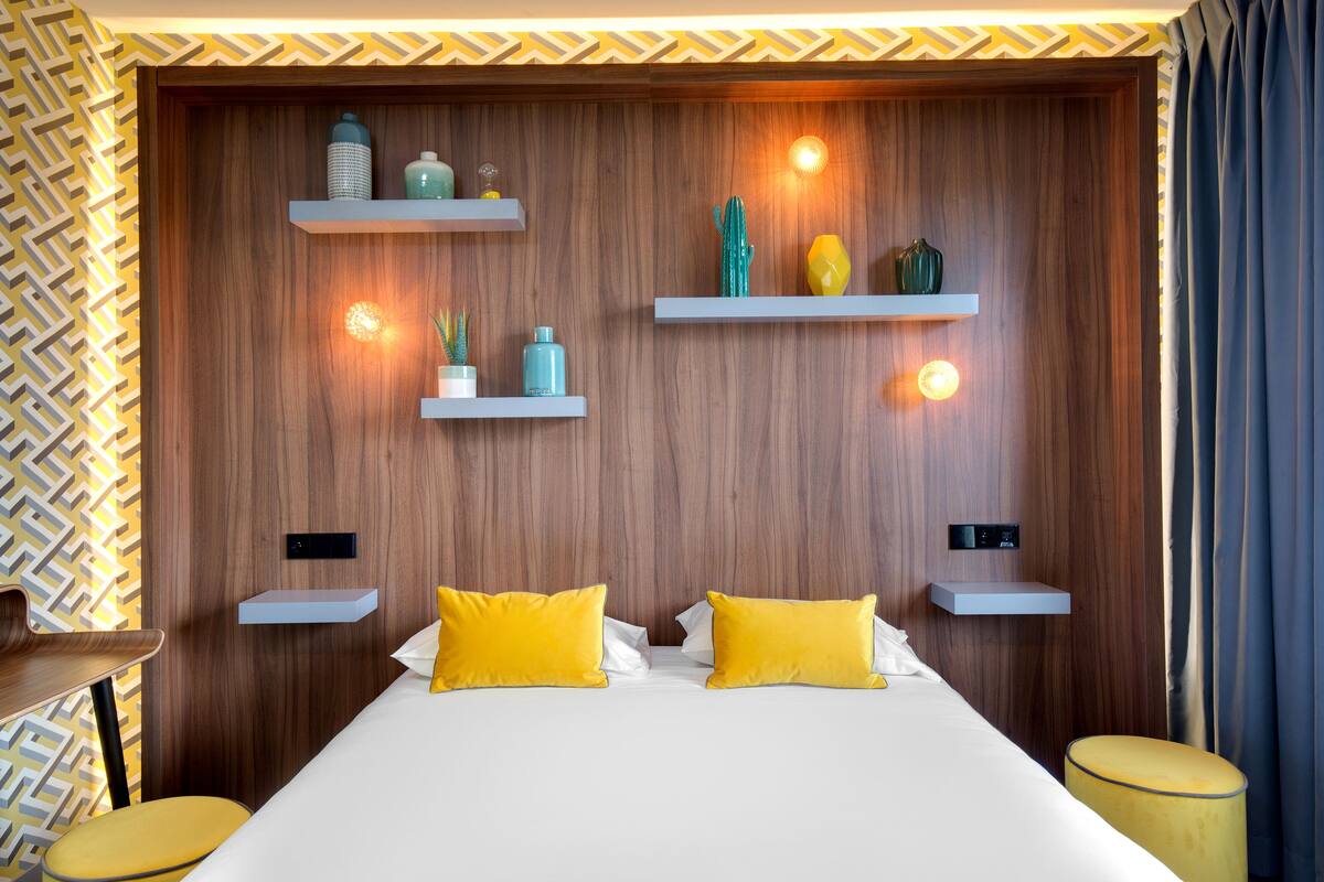 Ein modernes Hotelzimmer mit einem Holzkopfteil, zwei gelben Kissen und einem Bett mit weißer Bettwäsche. Es gibt zwei Nachttische mit kleinen Ablagen, die dekorative Gegenstände beherbergen.