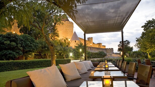 Un área de comedor al aire libre con mesas y sillas, una marquesina y un castillo al fondo.