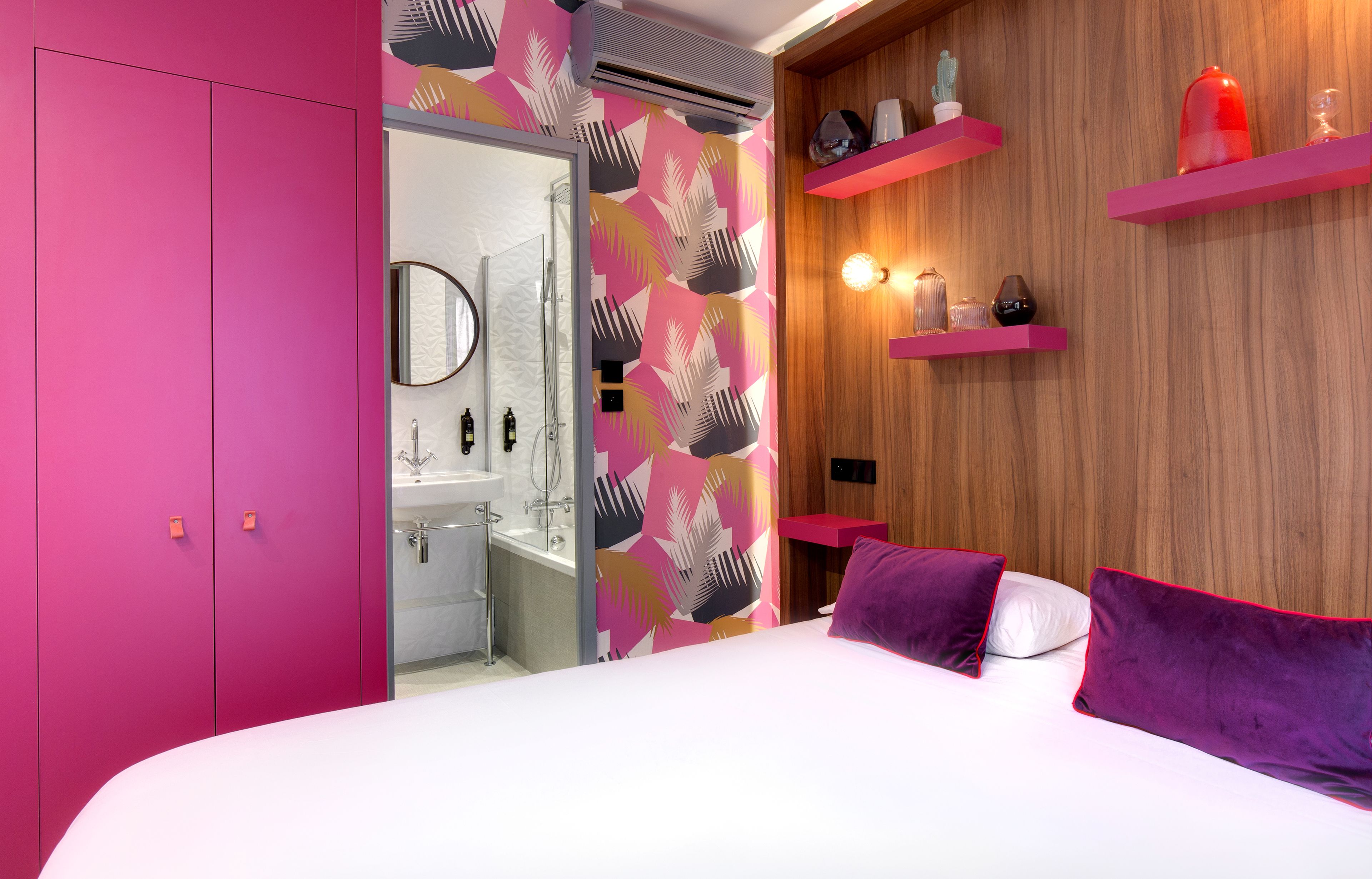 Camera da letto moderna con un letto grande, un armadio rosa, un bagno con doccia in vetro e mensole in legno con oggetti decorativi.