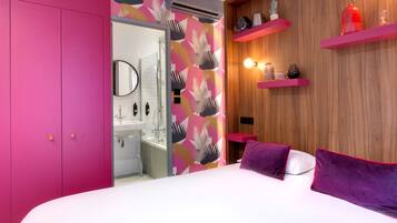 Un dormitorio moderno con una cama grande, un armario rosa, un baño con ducha de vidrio y estantes de madera con objetos decorativos.